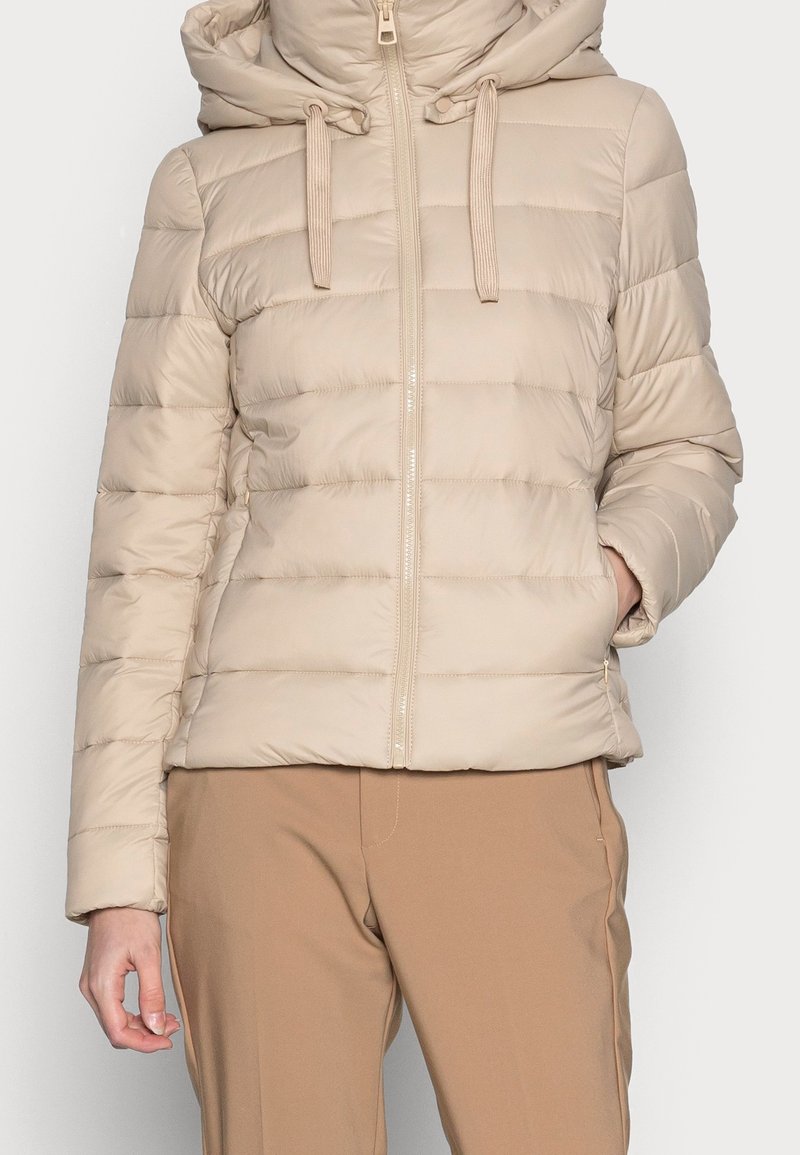 Doudoune matelassée beige avec capuche et fermeture éclair à l'avant, portée avec un pantalon marron clair, main dans la poche latérale, sur fond uni.