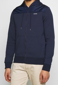 Sudadera con cremallera de color marino, fabricada en material suave, con capucha de satén, cordones ajustables, bolsillos delanteros y un logo en el pecho.