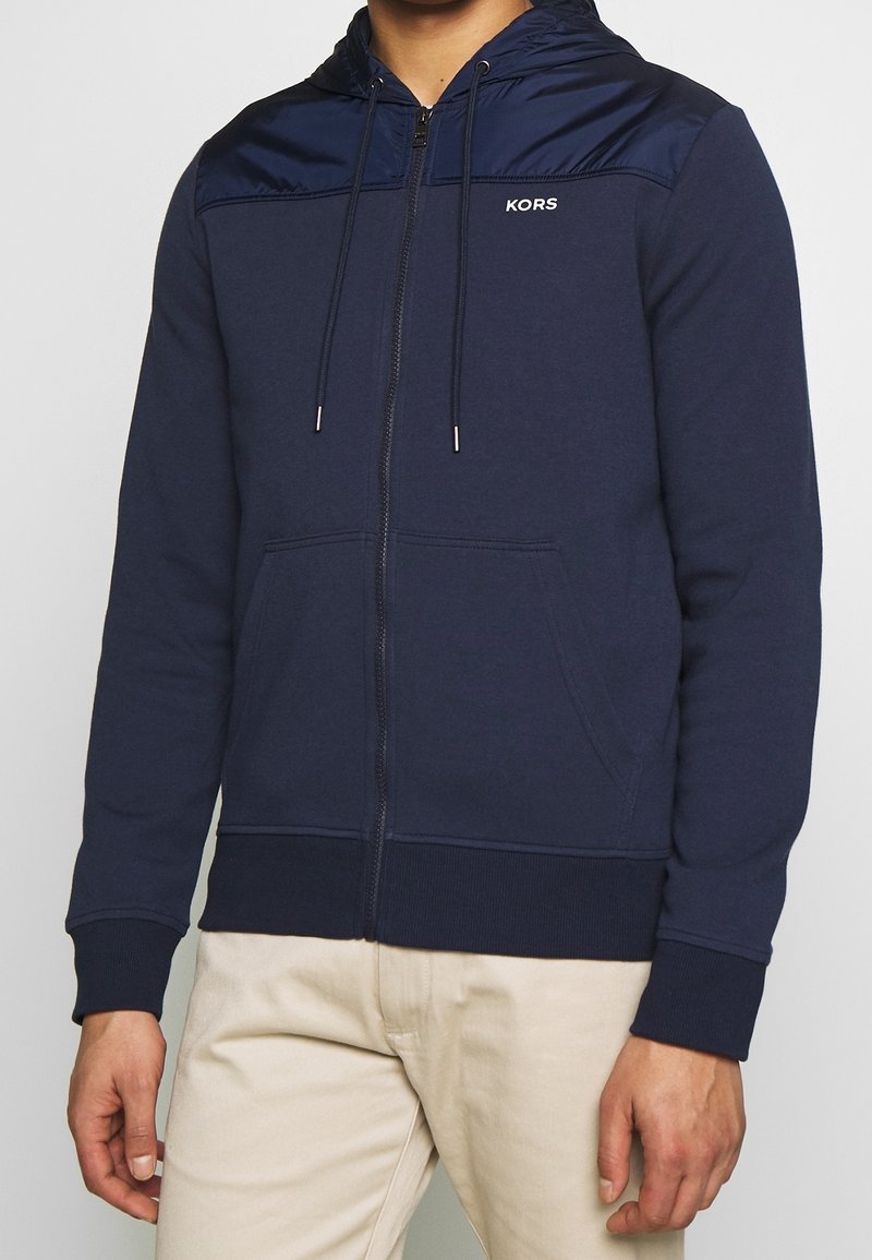 Sudadera con cremallera de color marino, fabricada en material suave, con capucha de satén, cordones ajustables, bolsillos delanteros y un logo en el pecho.