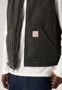 Σκούφο γκρί γιλέκο από υφή υφάσματος, με φερμουάρ στο μπροστινό μέρος και μία μεγάλη τσέπη με ετικέτα. Φοριέται πάνω από ένα λευκό ριμπ μπλουζάκι.