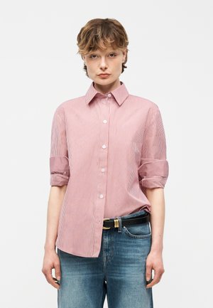 Rövid hajú fiatal visel egy piros-fehér csíkos, gombos, felhajtott ujjú inget és kék farmert fekete övvel.