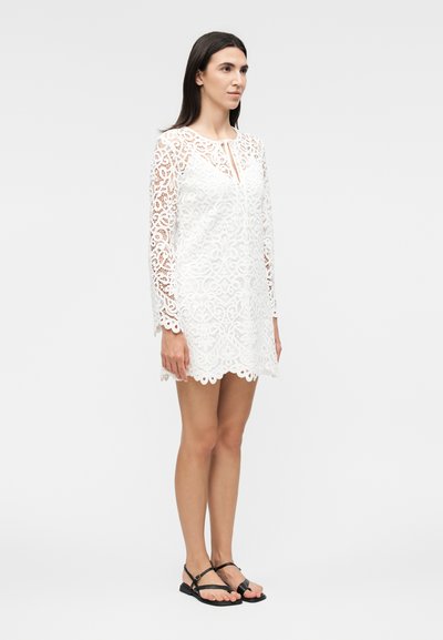 Femme debout portant une robe blanche en dentelle à manches longues avec des bords festonnés et des sandales plates noires, légèrement tournée vers la droite sur un fond uni.