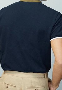 Mies, joka on pukeutunut tummansiniseen polo-paitaan, jossa on sinapin värinen kaulus ja vaaleat housut, nähtynä takaa neutraalilla taustalla.