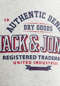 Pilkas medvilnės audinys su ryškiu, įvairiaspalviu tekstu: "AUTENTIŠKOS DENIMO SAUGOS PREKĖS JACK & JONES REGISTRUOTA PREKĖS ŽENKLAS VIENINGOS INDUSTRIJOS."