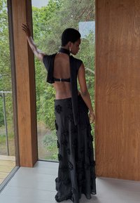 Zwart outfit met een rugloze ontwerpen en een halslijn met strik. Lange rok versierd met textuur zwart bloem appliqués. Achtergrond van houten panelen.