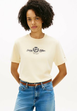 Femme aux cheveux bouclés portant un t-shirt crème Tommy Hilfiger et un jean bleu avec une ceinture marron, debout devant un fond blanc uni.