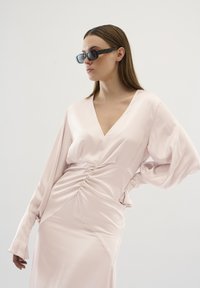 Rochie din satin roz deschis, cu decolteu în V, mâneci lungi și detalii plisate la talie. Asociată cu ochelari de soare negri, dreptunghiulari.