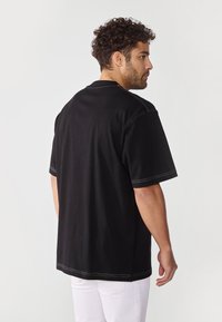 Sort T-shirt med korte ærmer og synlige hvide syninger. Løs pasform, rund halsudskæring og blødt stof. Bæres med hvide bukser.