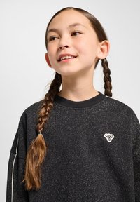 Svart sweatshirt med glittrande textur, rund halsringning och vita kontraststripes. Har en broderad ljusfärgad logga på bröstet.