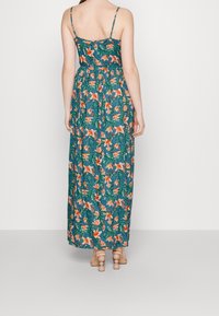 Kvinna i en lång teal klänning med orange blommönster och spaghettiband, ståendes barfota mot en helt vit bakgrund.