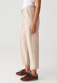 Vue de côté d'une personne portant un pantalon beige à longueur de cheville avec taille à cordon et des baskets marron à lacets sur fond clair.