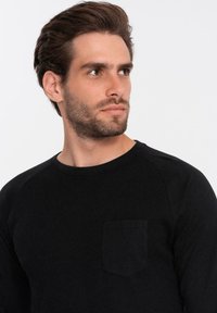 Mustapaita, jossa on pitkät hihat, ribbaustyyppinen pinta, pyöreä kaula-aukko ja pieni tasku vasemmassa rinnassa. Hihnat ovat raglan-tyyppiset.