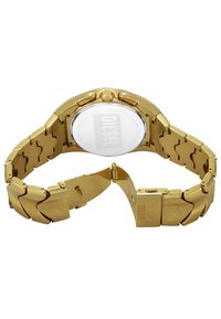 Montre en acier inoxydable doré avec un bracelet à maillons, boîtier rond et un fond transparent affichant le logo Diesel. Fini lisse et poli.