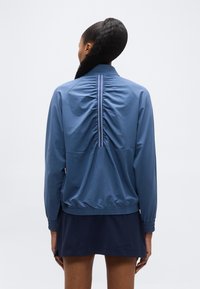 Γυναίκα με μακριά μαλ λιά δεμένα πίσω, φοράει μπλε μπουφάν με ραφή με πιέτες και αντανακλαστική λωρίδα στην πλάτη, συνδυασμένο με μπλε σκούρο φούστα.