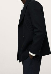 Granatowa wełniana marynarka dwurzędowa z dwoma przednimi kieszeniami i czarnymi guzikami. Ma klapowany kołnierz i widoczną białą mankietę na nadgarstku.