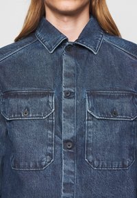 Chaqueta de mezclilla en lavado azul oscuro, con cuello puntiagudo, cierre de botones y dos bolsillos con solapa con botones negros.