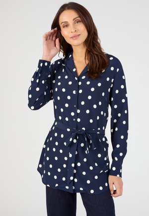 Mujer vistiendo una blusa azul marino de manga larga con botones y lunares blancos, con un cinturón anudado en la cintura, posando con una mano cerca del rostro.