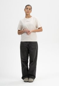 Beige Baumwoll-T-Shirt mit kurzen Ärmeln und Rundhalsausschnitt; mit einem kleinen schwarzen Textdetail auf der Brust. Getragen mit dunklen Jeans.