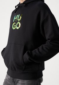 Sudadera negra con bolsillo delantero, que presenta un logo reflectante de "HUGO" en verde y dorado. Capucha con cordones y puños acanalados.