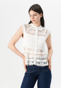 Blusa bianca senza maniche con dettagli in pizzo, chiusura a bottoni e orlo scallop. Il tessuto presenta intricate fantasie floreali e strisce testurizzate.