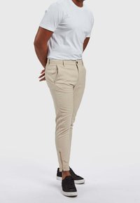 Mężczyzna w beżowych, dopasowanych spodniach z suwakiem przy kostkach, białej koszulce i czarnych trampkach stoi z rękami za plecami na białym tle.