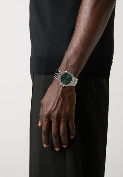 Stříbrné kovové hodinky se zeleným ciferníkem, s elegantním tvarem a vzájemně propojeným páskem. Noseny na ruce s tmavou pletí na černém pozadí.