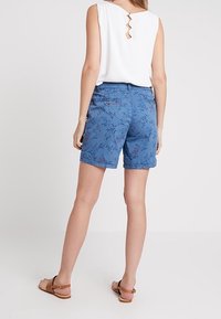 Shorts bleu à motifs floraux en tissu léger, dotés d'une poche arrière, associés à un haut sans manches blanc avec un dos découpé.