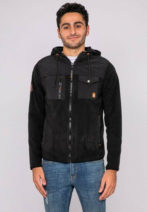 Jeune homme aux cheveux bouclés foncés portant une veste noire à fermeture éclair avec capuche et multiples poches, debout devant un fond clair uni.