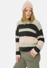 Vrouw in een roze gebreide beanie, gestreepte zwarte, roze en grijze trui, en olijfgroene broek met één hand in haar zak.