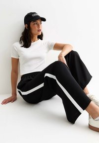 Svarta träningsbyxor med vita sidostråk, kombinerade med en åtsittande vit t-shirt och en svart basebollkeps. Skorna är vita sneakers.