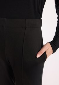 Czarne dopasowane spodnie o gładkiej strukturze, elastycznym pasku w talii i kieszeniach bocznych. Detale obejmują subtelną szew wzdłuż długości spodni.