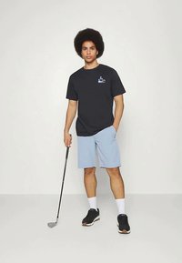 Mustapaitainen lyhythihainen t-paita logolla, vaaleansiniset shortsit, mustat lenkkarit ja harmaa golfmaila yhtenäisellä taustalla.