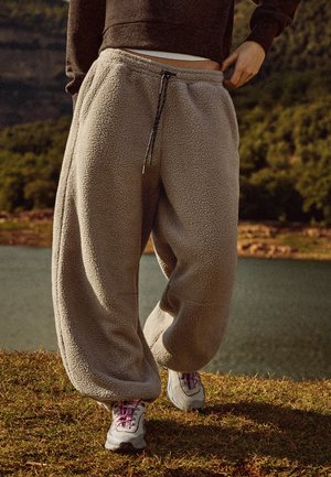Une personne en extérieur ajuste la taille d’un pantalon de jogging beige en polaire. Elle porte un haut foncé et des baskets grises à lacets roses.