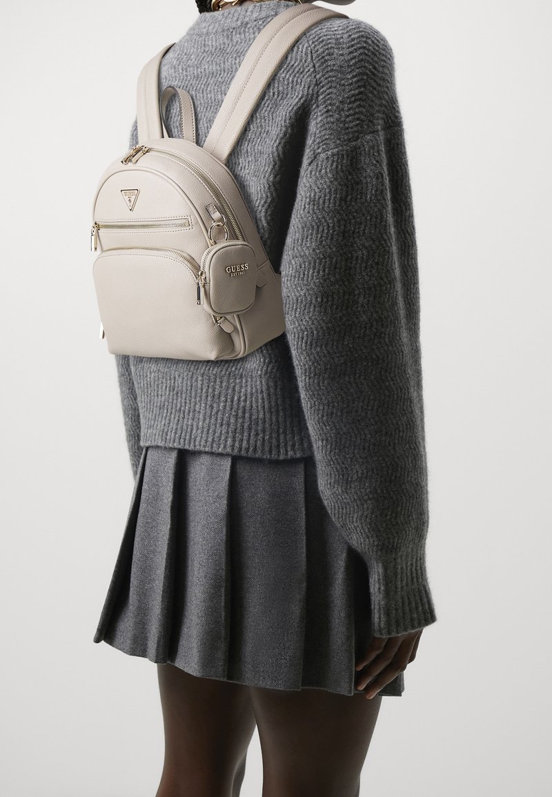 Sac à dos en cuir beige avec deux zips sur le devant, accent logo, bretelles ajustables et texture lisse, assorti à un sweater gris et une jupe plissée.