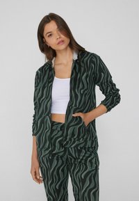 Mujer con chaqueta de cremallera a rayas de cebra verde y negra y pantalones a juego, con un top corto blanco, de pie frente a un fondo liso.