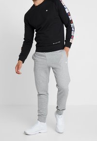 Felpa nera con logo sul petto e maniche a fantasia, abbinata a pantaloni della tuta grigi e sneakers bianche. Abbigliamento sportivo casual.