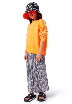Sweatshirt orange, jupe maxi noire et blanche à motifs, chapeau seau noir avec texte blanc, et sandales noires à glissière avec logo blanc.