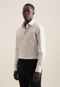 Chemise blanche à boutonnières avec de petits pois noirs, manches longues et col. Présente des boutons dorés et une coupe ajustée. Portée avec un pantalon noir.