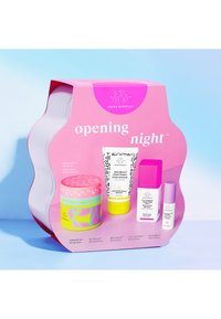 Pink packaging featuring a wavy design holds four skincare products: a colorful jar, a white tube, and two smaller bottles, all with distinct labels.