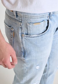 Svijetloplave traperice s ispucanim dizajnom, koje sadrže vidljive rupice i patentni zatvarač. Imaju više džepova i detalj u boji zemlje.