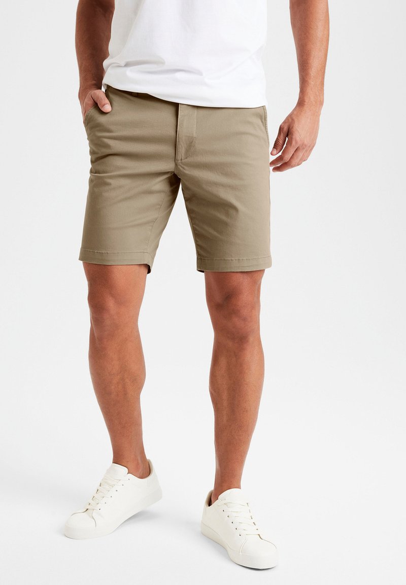 Pantaloni corti khaki realizzati in tessuto misto cotone, con vita piana, tasche laterali e una vestibilità comoda, abbinati a sneaker bianche.