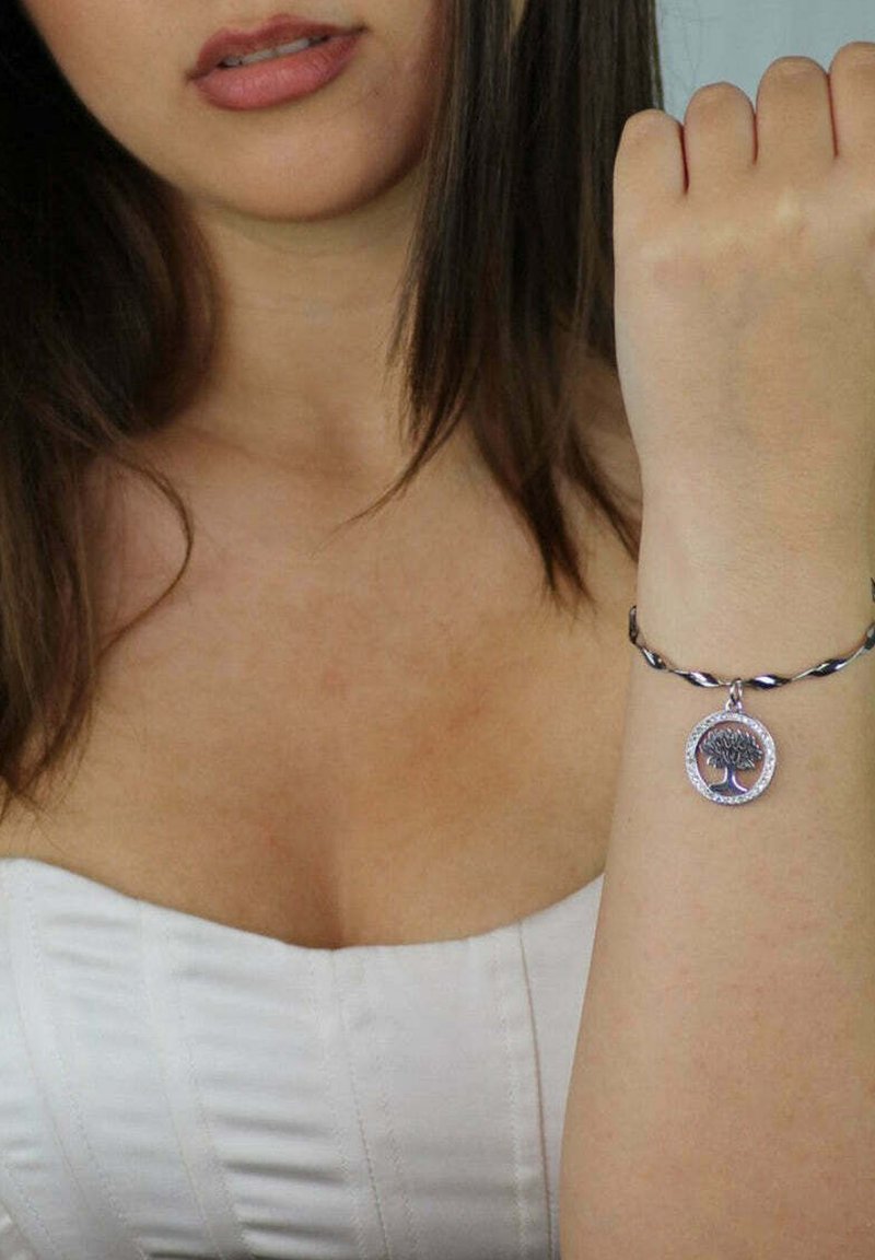 Bracciale d'argento con un design intrecciato, caratterizzato da un charm circolare raffigurante un albero all'interno di un bordo decorativo. Indossato su un polso di pelle chiara.