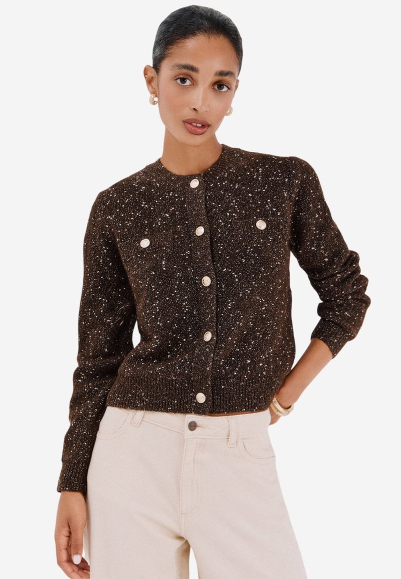 Cardigan marrone con texture e macchie bianche, colletto rotondo e bottoni frontali. Ha maniche lunghe e due piccoli taschini frontali.
