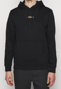 Sudadera negra con capucha en tela suave con un bolsillo tipo canguro, que presenta un emblema dorado en el pecho y cordones ajustables.