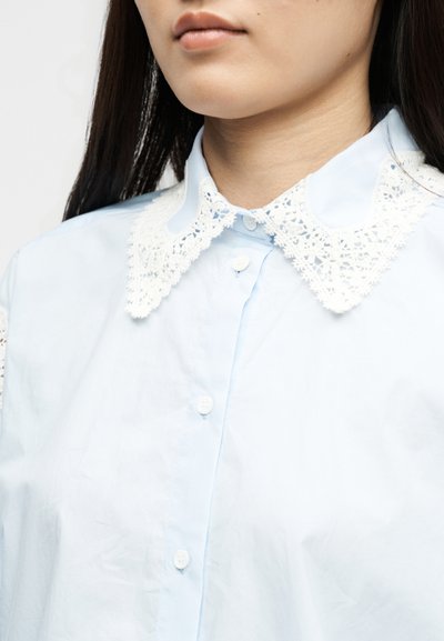Κοντινό πλάνο ενός ατόμου που φορά ένα ανοιχτό μπλε πουκάμισο με λευκό δαντελωτό γιακά και έχει μακριά σκούρα μαλλιά ορατά.