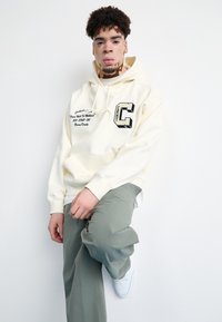 Sudadera crema con bolsillo frontal, diseño gráfico negro y detalles bordados; combinada con pantalones verdes holgados y zapatillas blancas.