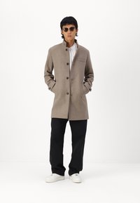 Beige ullfrakk med stående krage, frontknapper og brystlomme; kombinert med svarte bukser og hvite joggesko. Modellen har solbriller på.
