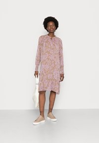 Vestido de manga longa em castanho claro com um padrão floral lilás, apresentando um decote franzido e uma bainha com volantes, combinado com ténis bege.