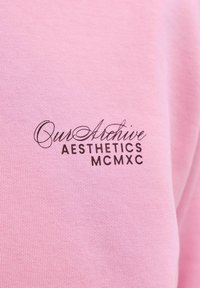 Hele roosa puuvillane dressipluus, millel on musta trükitud tekst "Our Archive AESTHETICS MCMXC" käsikirjas ja paksus kirjas. Sile tekstuur.