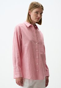 Gestreifte Button-Up-Bluse aus roter und weißer Baumwolle mit Kragen, langen Ärmeln und einer Brusttasche. Weiche Textur und lockerer Schnitt.